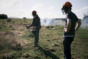 One person using a brush cutter with another looking on with a bonfire in the background